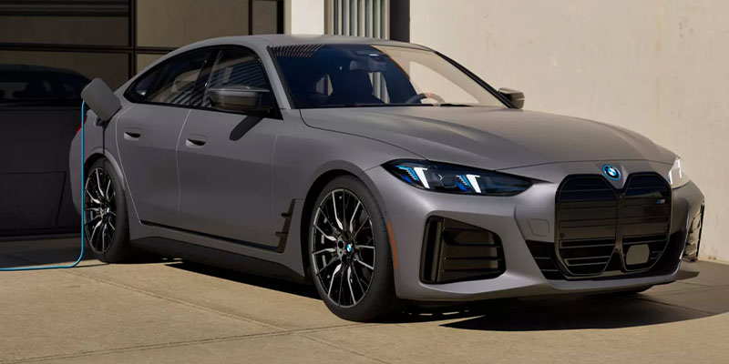 2025 BMW i4 electric car parked, charging cable attached, sleek gray design, sporty black wheels.